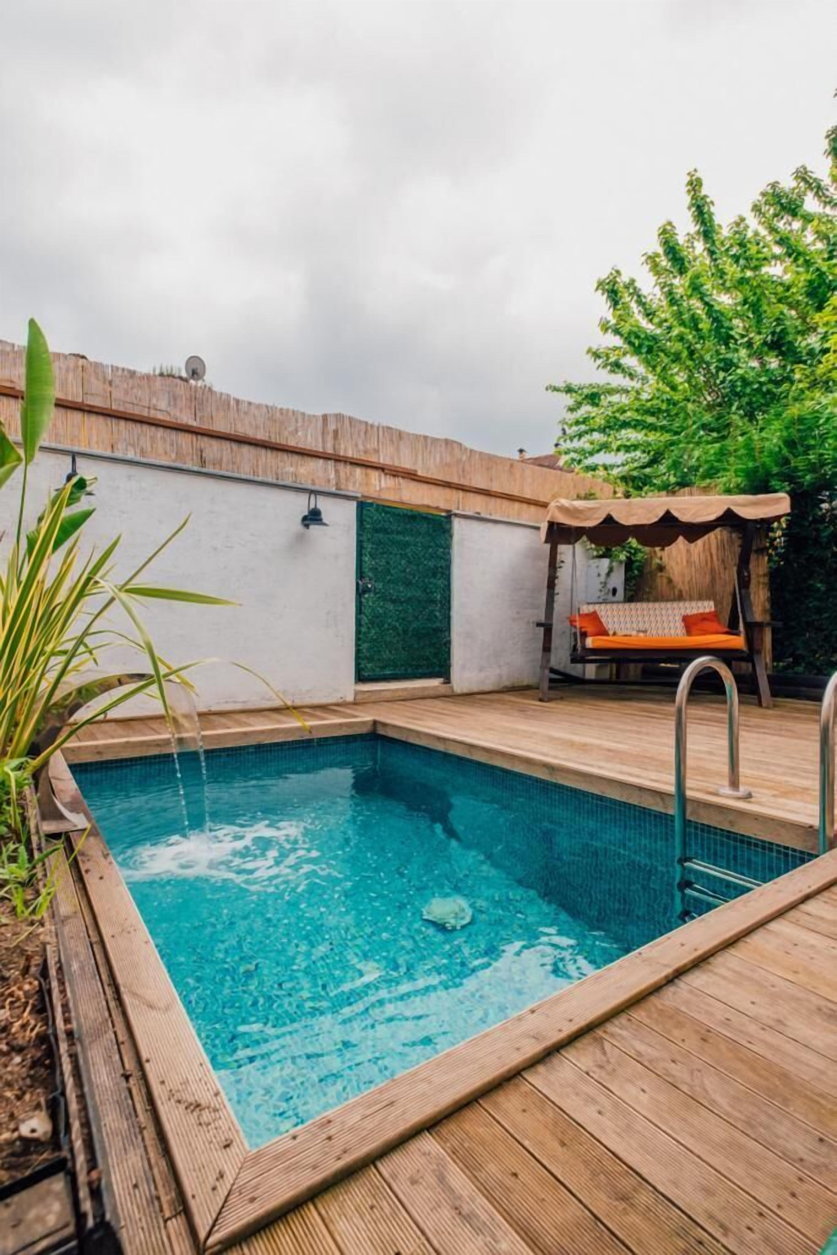 A rectangular pool with a wooden deck, a swing chair, and a green wall.