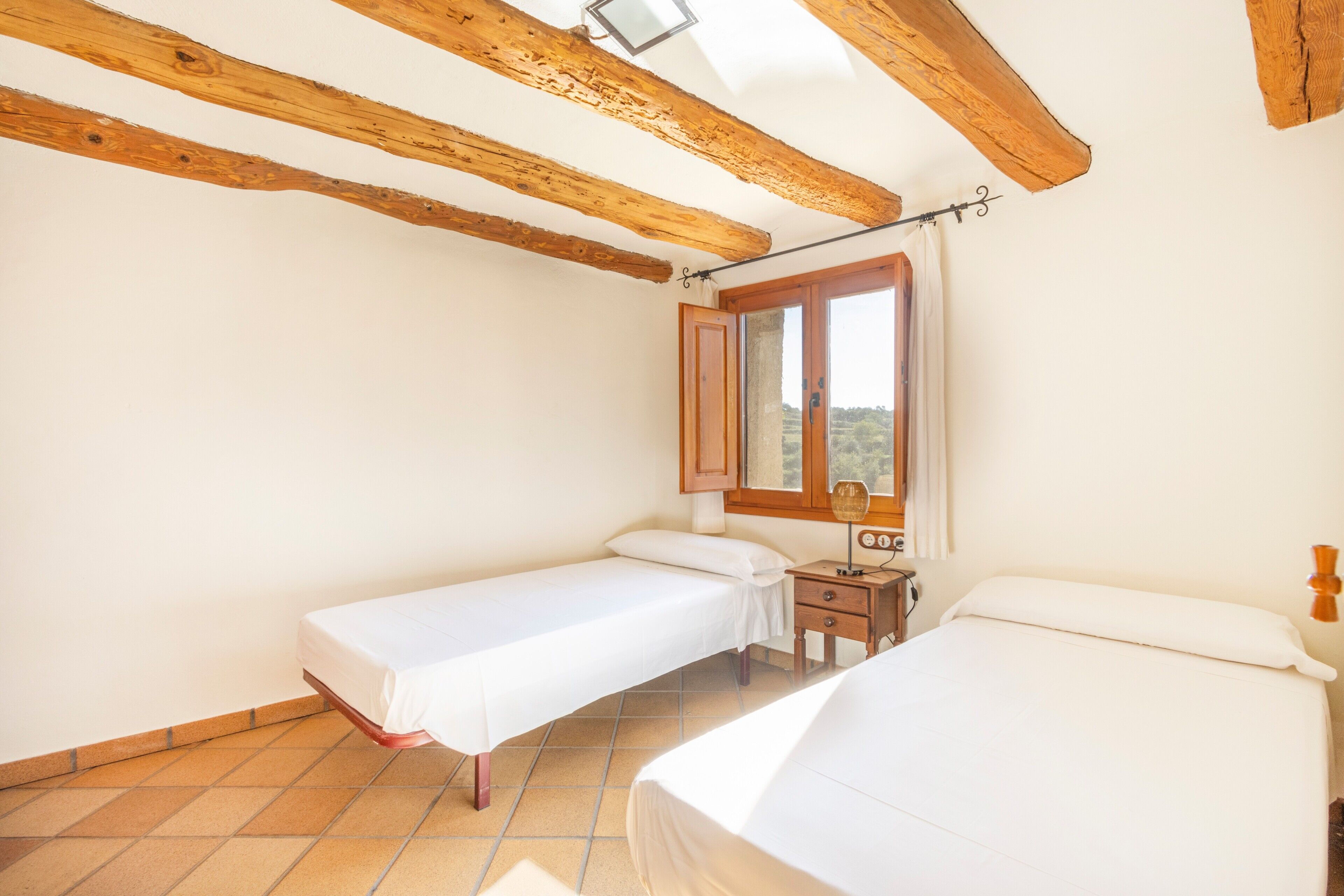 Un dormitorio con dos camas, vigas de madera en el techo, una ventana con cortinas y una mesita de noche.