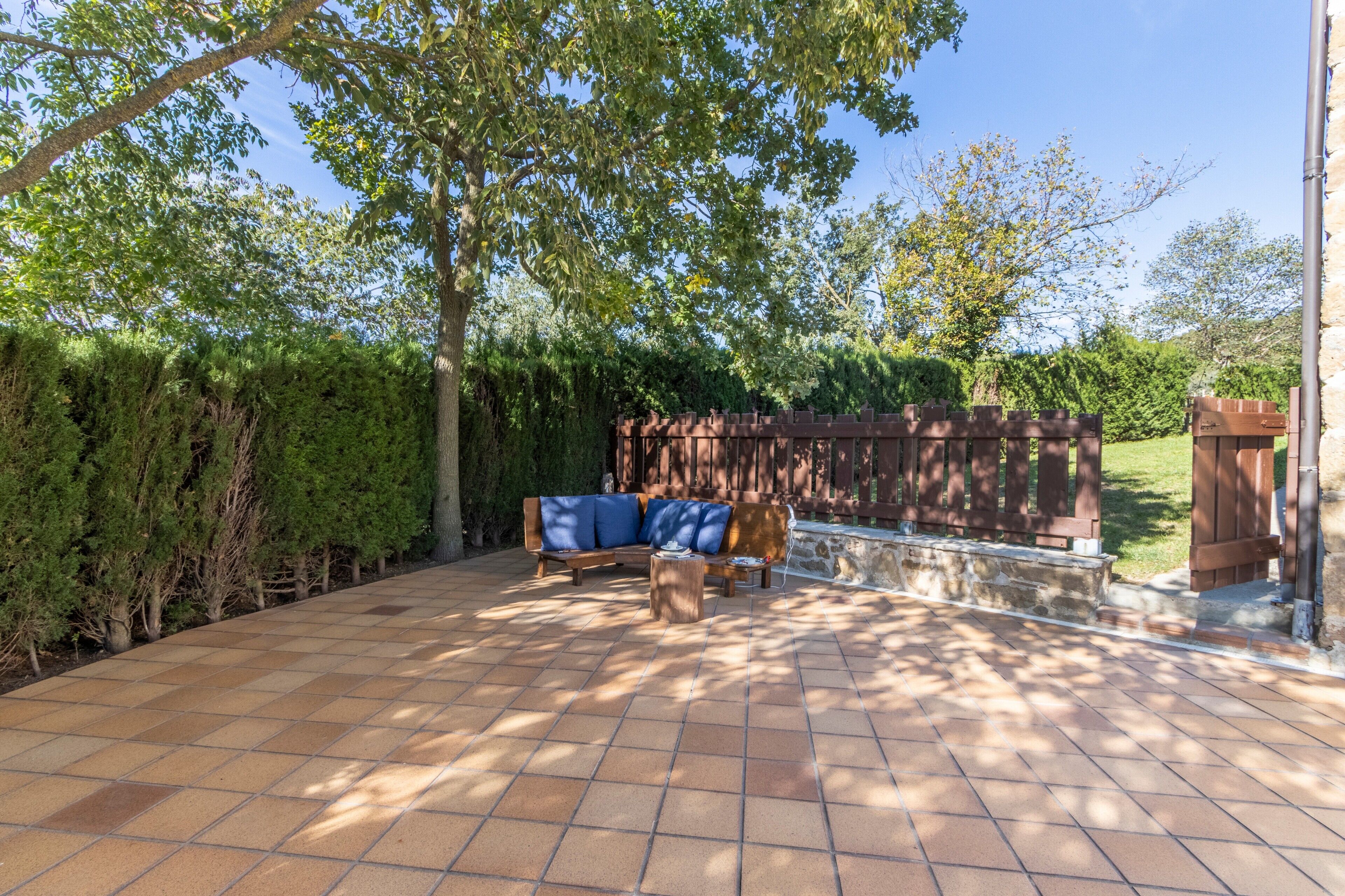 Un patio con un banco de madera y un sofá, rodeado por un seto y árboles.