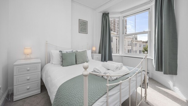 Et soveværelse med en hvid seng, et natbord med lampe, en kommode og et vindue med gardiner.