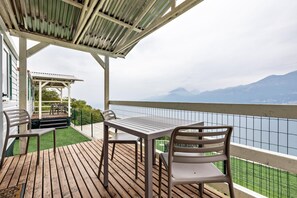 A wooden deck with a table and chairs, overlooking a body of water and mountains.