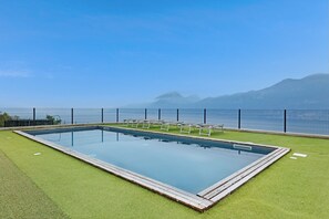 A rectangular swimming pool with a wooden deck, surrounded by artificial grass, and a fence with chairs.