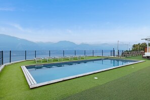 Une piscine rectangulaire dotée d’une terrasse en bois, entourée de gazon synthétique, et offrant une vue sur les montagnes et la mer.