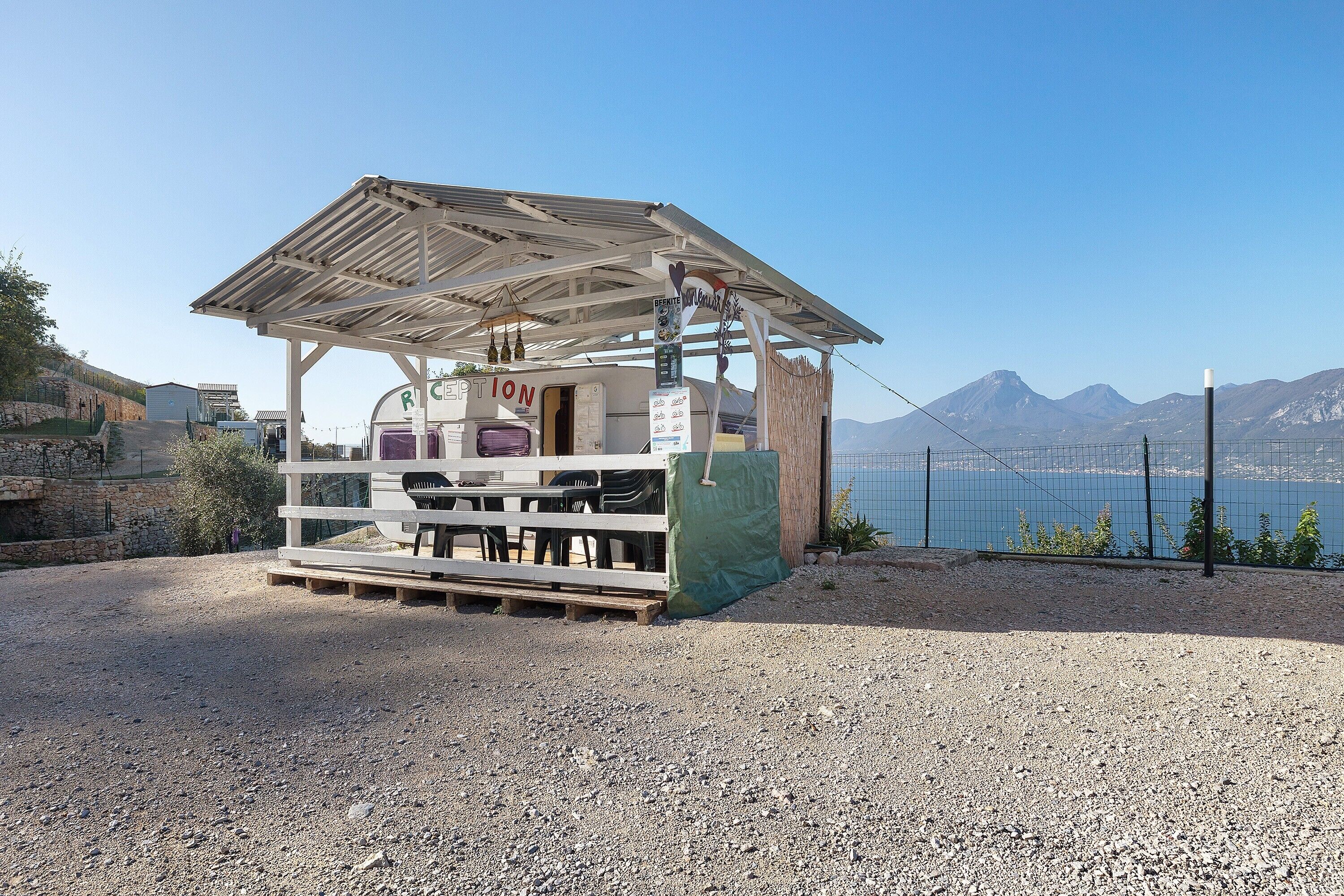 Un pequeño quiosco al aire libre con techo metálico, una mesa con sillas y un cartel que dice 'RECEPTION'.