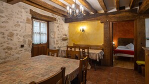Una habitación rústica con vigas de madera a la vista, una pared de piedra, una mesa de comedor con mantel estampado y una cama con colcha roja y blanca.
