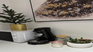 Una encimera de cocina con una cafetera, una pequeña planta en maceta, un tarro y un cuadro en la pared.
