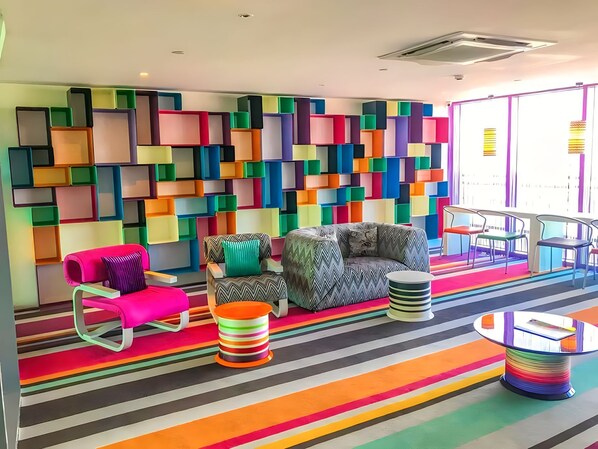 A vibrant room with colorful geometric wall panels, striped carpet, and modern furniture including a sofa, armchair, and ottoman.