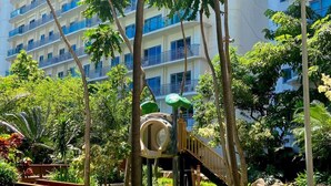 Un parque infantil con sube y bajas y un tobogán, rodeado de abundante follaje y un edificio de varios pisos con balcones.