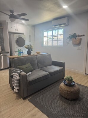 A modern living room with a grey sofa, a ceiling fan, an air conditioning unit, a window with a view, and a kitchen area with a washing machine and a sink. - Cozy Guesthouse w/Full Kitchen in Oxnard (Oxnard)