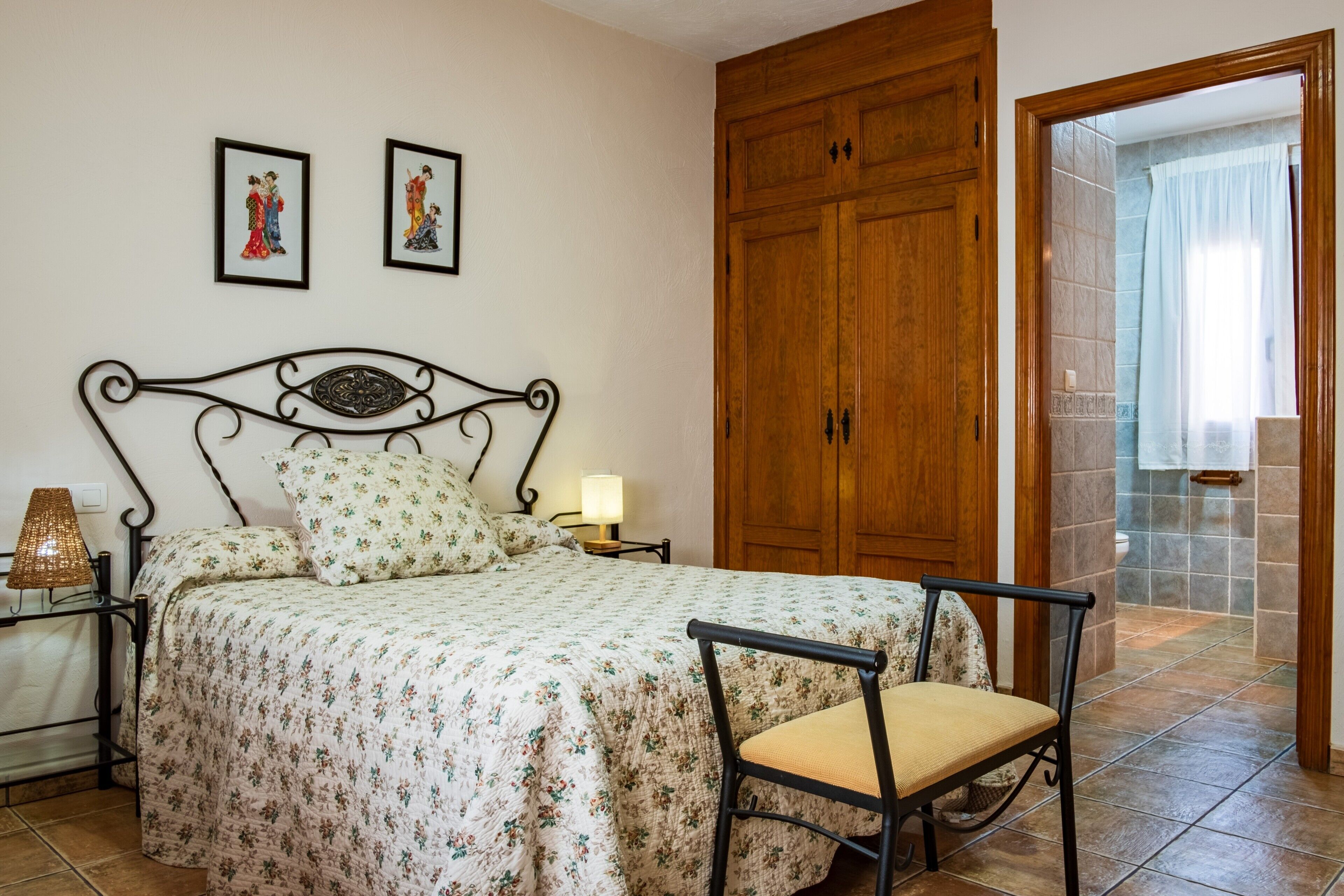 Un dormitorio con cama, una silla, una mesita de noche, lámpara, armario de madera y dos cuadros en la pared.