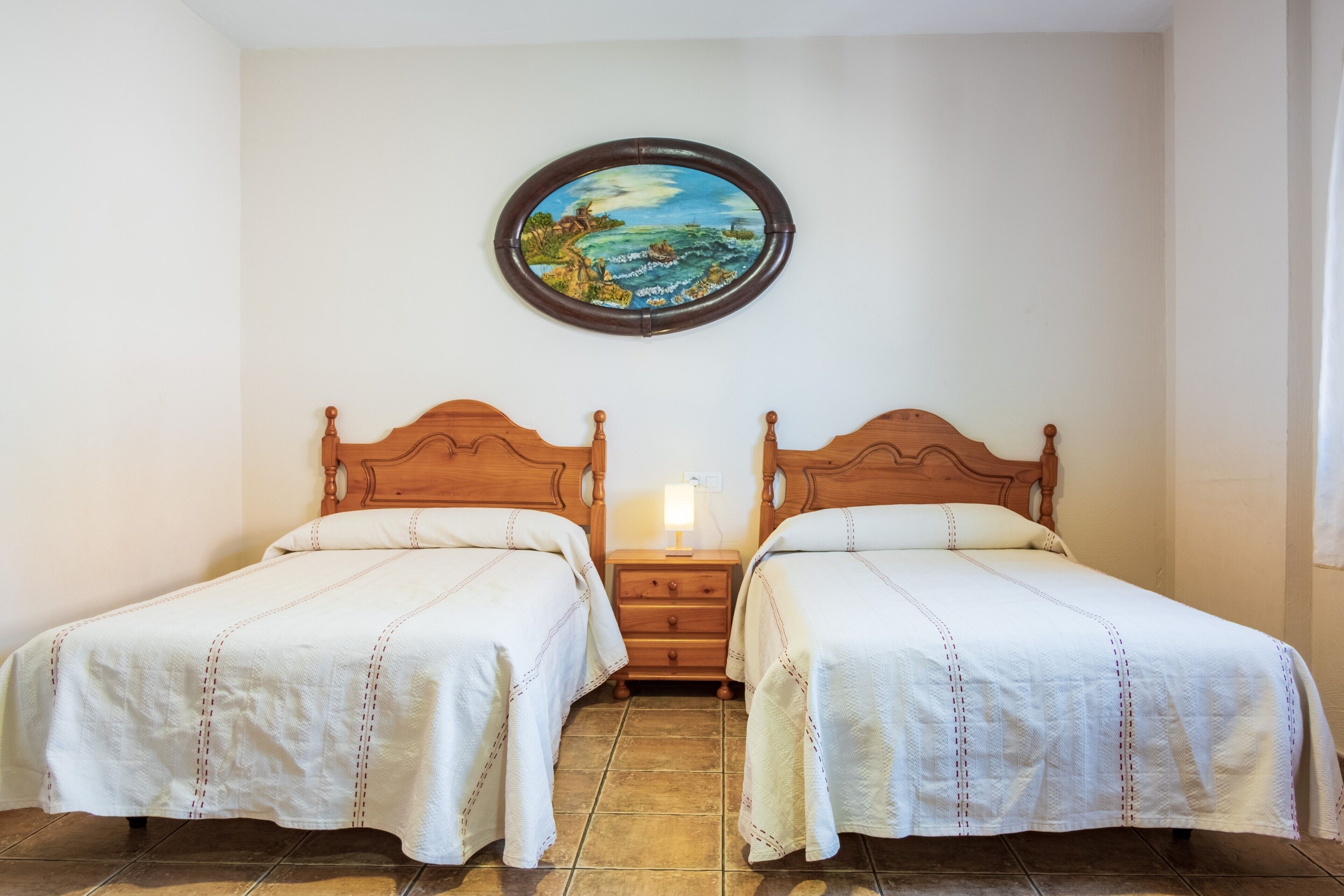 A room with two wooden beds, a nightstand, a wall-mounted painting, and a tiled floor.
