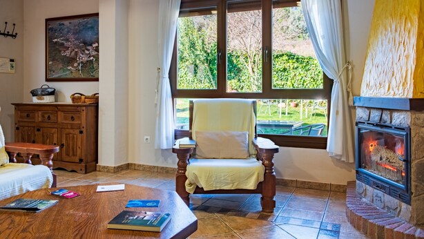 Une pièce chaleureuse avec une cheminée, une chaise en bois, une table avec des livres et une fenêtre avec des rideaux.