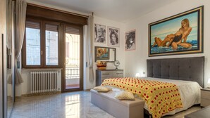 Un dormitorio con una cama grande, una ventana con cortinas, un radiador, una mesita de noche y un cuadro en la pared.