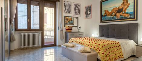 Un dormitorio con una cama grande, una ventana con cortinas, un radiador, una mesita de noche y un cuadro en la pared.