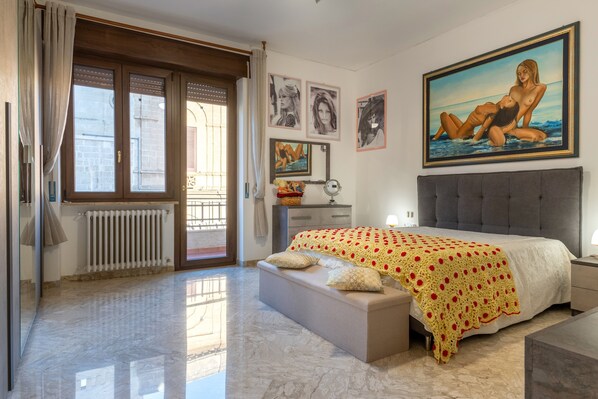 A bedroom with a large bed, a window with curtains, a radiator, a bedside table, and a painting on the wall.