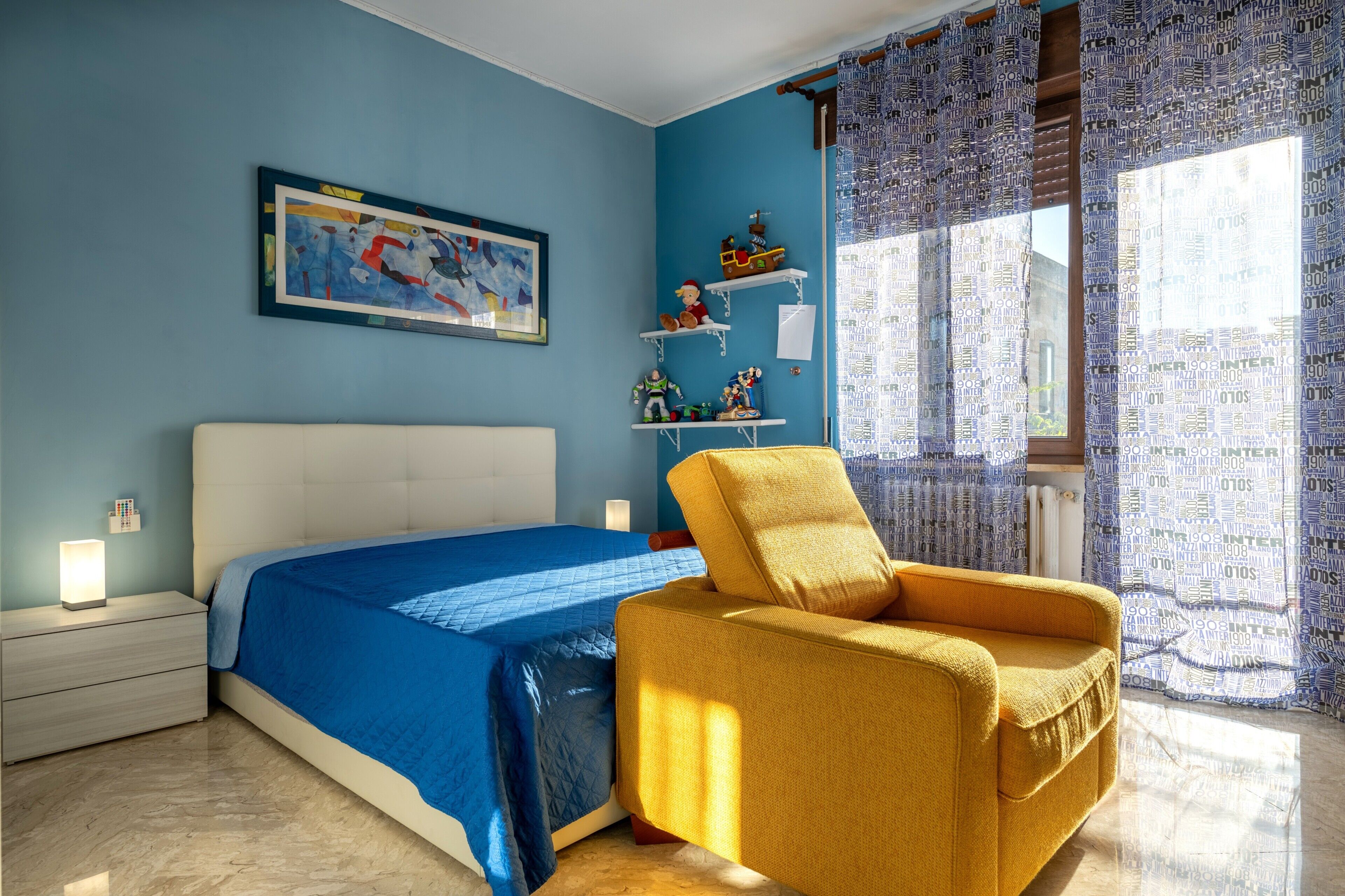 Una camera da letto con un letto, una sedia, un comodino, uno scaffale con giocattoli e una finestra con tende.