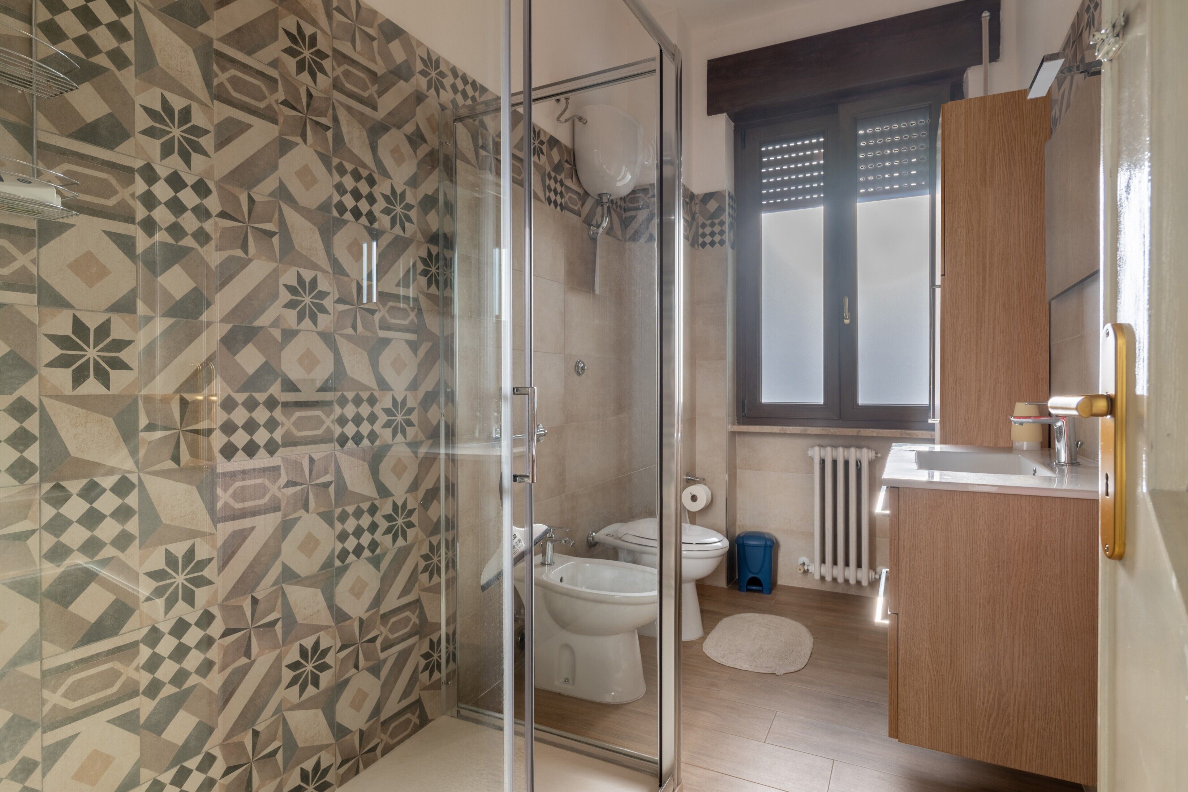 A bathroom with patterned tiles, a glass shower enclosure, a toilet, and a sink with a wooden cabinet.