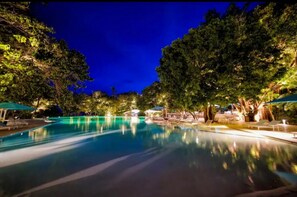 Une piscine nocturne, dotée de chaises longues et de parasols, entourée d’arbres.
