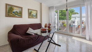 Una habitación con un sofá marrón, una mesa de vidrio, dos cuadros enmarcados y vistas a un jardín a través de amplias ventanas.