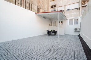 Ein Innenhof mit Fliesenboden, einer überdachten Sitzgelegenheit mit Stühlen und Tisch sowie ein Gebäude mit Balkon.