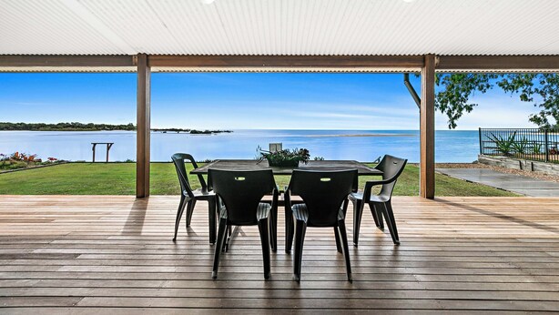 Ein überdachter Außenbereich mit Holzterrasse, einem runden Tisch mit Stühlen und Blick auf ein Gewässer.