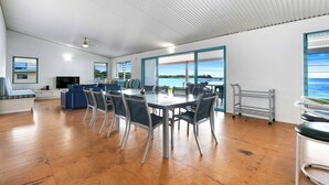 Ein geräumiges Zimmer mit einem großen Esstisch, Stühlen, einer Couch und einem Fernseher. Große Fenster bieten Blick aufs Meer.