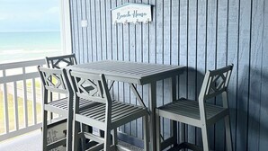 Een houten terras met een tafel en stoelen, een bord met de tekst 'Welkom in Ons Strandhuis' en uitzicht op de oceaan.
