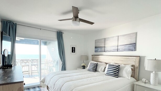 Een ruime slaapkamer met een groot bed, houten vloeren, een plafondventilator en uitzicht op de oceaan door het raam.