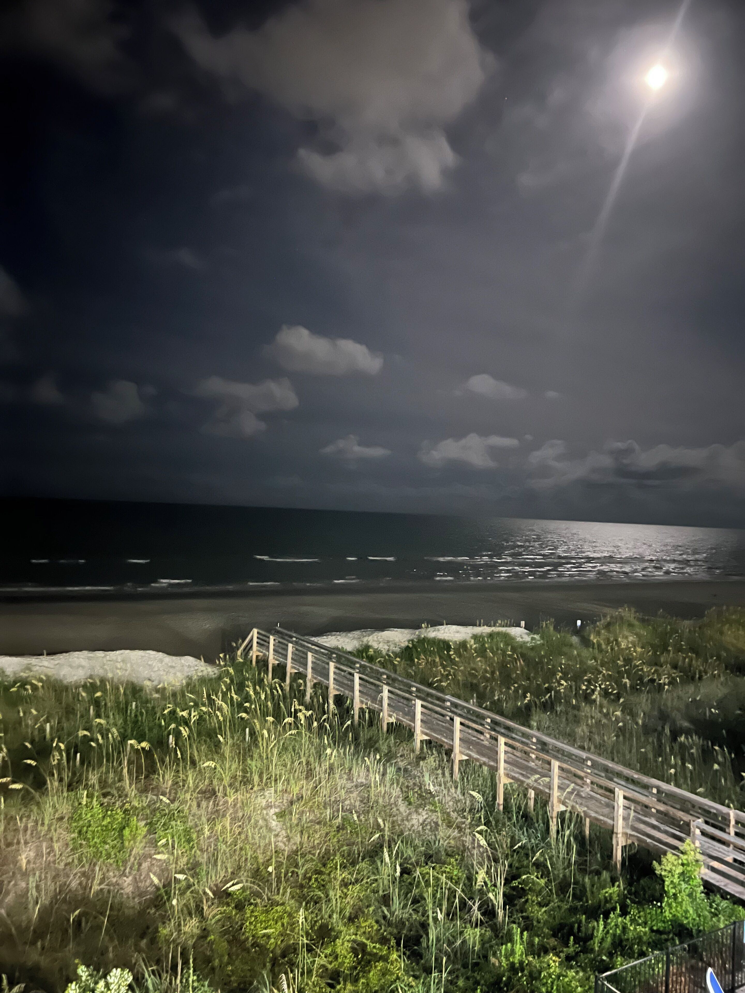 A nighttime coastal scene with a wooden staircase leading down to the beach, under a cloudy sky with a visible moon.