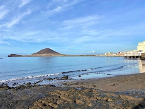 Paesaggio costiero con spiaggia, costa rocciosa, una montagna e edifici sullo sfondo.