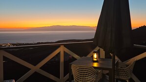 Een balkon met een tafel en stoelen, met uitzicht op een kustlandschap tijdens zonsondergang.