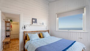 Een slaapkamer met een houten bed, een raam met uitzicht, een plank met spullen, en een deur die naar een andere kamer leidt.