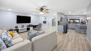 Ein modernes Wohnzimmer mit Flachbildfernseher, Deckenventilator und Essbereich, mit einer Küche im Hintergrund.