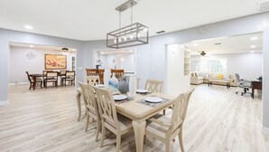 Ein geräumiger Essbereich mit Holztisch und Stühlen, einer modernen Kronleuchter und Blick in ein Wohnzimmer mit Sofa und Schreibtisch.