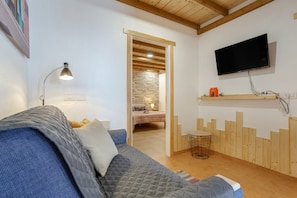 A room with a bed, a TV mounted on the wall, a wooden shelf, and a door leading to another room.