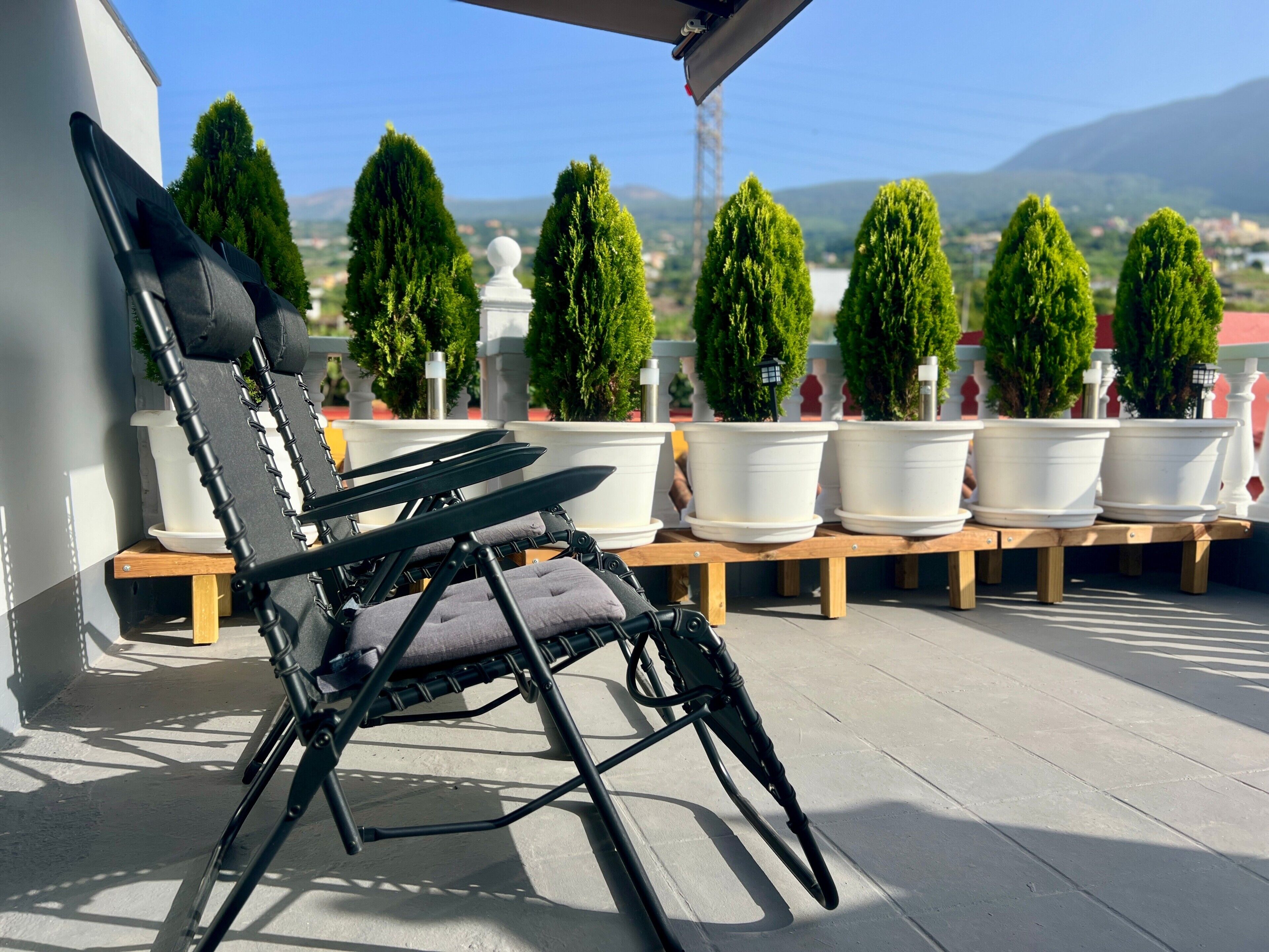 A black lounge chair with a cushion, positioned on a wooden bench. In the background, there are several potted plants and a clear view of a landscape with hills.