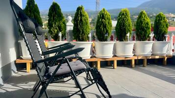 A black lounge chair with a cushion, positioned on a wooden bench. In the background, there are several potted plants and a clear view of a landscape with hills.