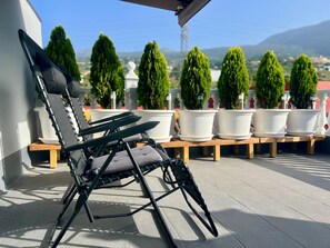 A black lounge chair with a cushion, positioned on a wooden bench. In the background, there are several potted plants and a clear view of a landscape with hills.