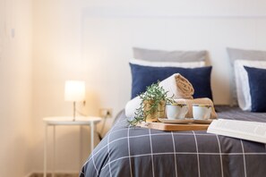 Una cama bien hecha con una colcha de cuadros, una toalla enrollada, dos tazas y una bandeja con una planta.