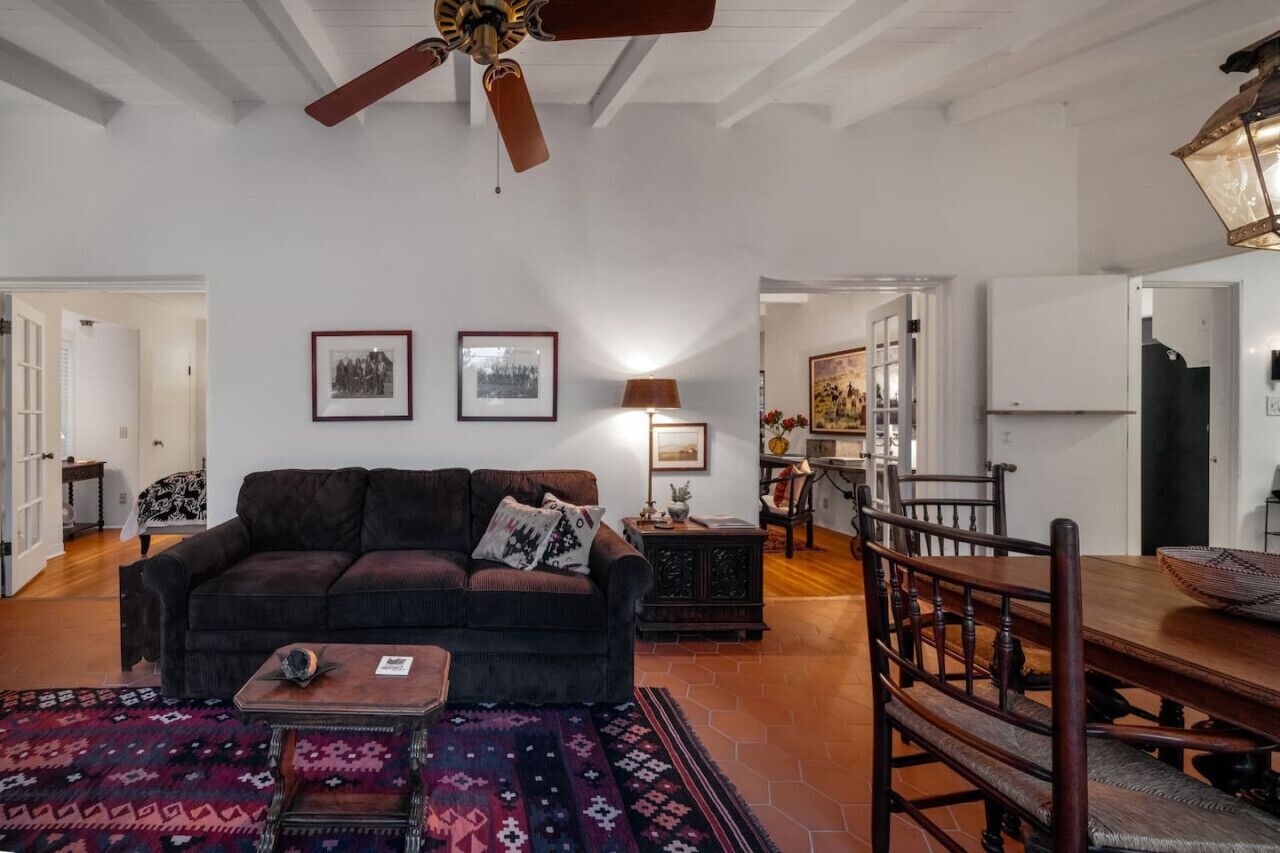 Uma sala de estar ampla com um sofá marrom escuro, um tapete estampado, uma mesa de centro de madeira e uma área de jantar com mesa e cadeiras de madeira.