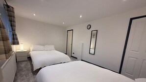 Habitación de hotel con dos camas individuales, una mesita de noche con lámpara, un reloj en la pared y una puerta.
