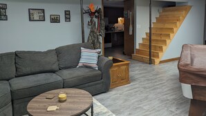 Un salon avec un canapé gris, une table basse en bois et un coussin à rayures. Un meuble en bois est adossé au mur, et un escalier mène à une autre pièce.