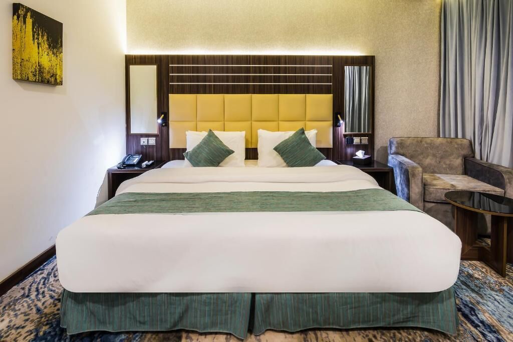 Une chambre d’hôtel avec un grand lit, une tête de lit ornée de lignes horizontales, une table de chevet avec une lampe, une chaise et une petite table.