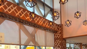 A spacious lobby with a large window, a brown sofa, a coffee table, and hanging geometric light fixtures. - White Diamond Hotel Sari (Jeddah)