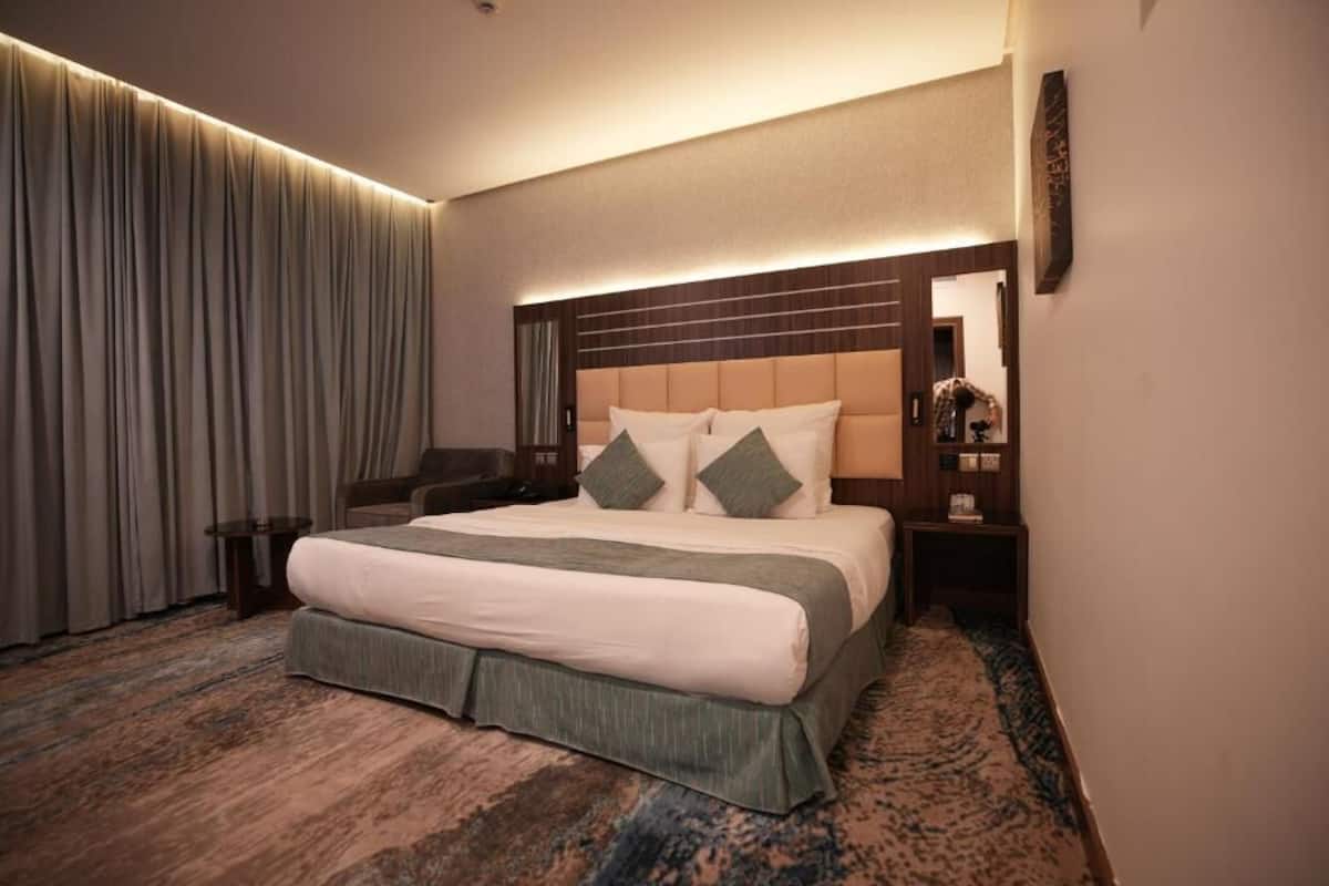 Habitación de hotel con una cama grande, cabecera con líneas horizontales, mesita de noche, escritorio y un ventanal con cortinas.