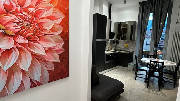 A modern living room with a large floral painting, a dark sofa, a dining area with a table and chairs, and a kitchen in the background.