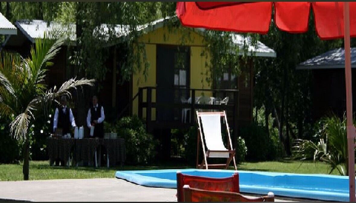 A pool area with a lounge chair and a small table, surrounded by greenery and a building with a yellow facade.