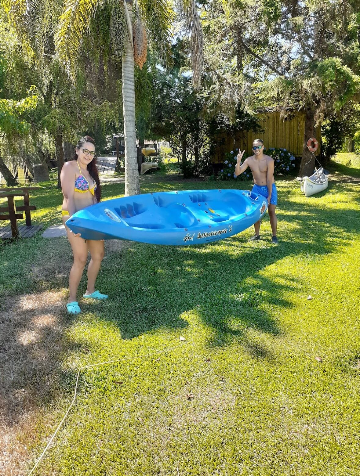 Dos personas sostienen una balsa inflable azul en un patio trasero con césped, árboles y una estructura de madera al fondo.