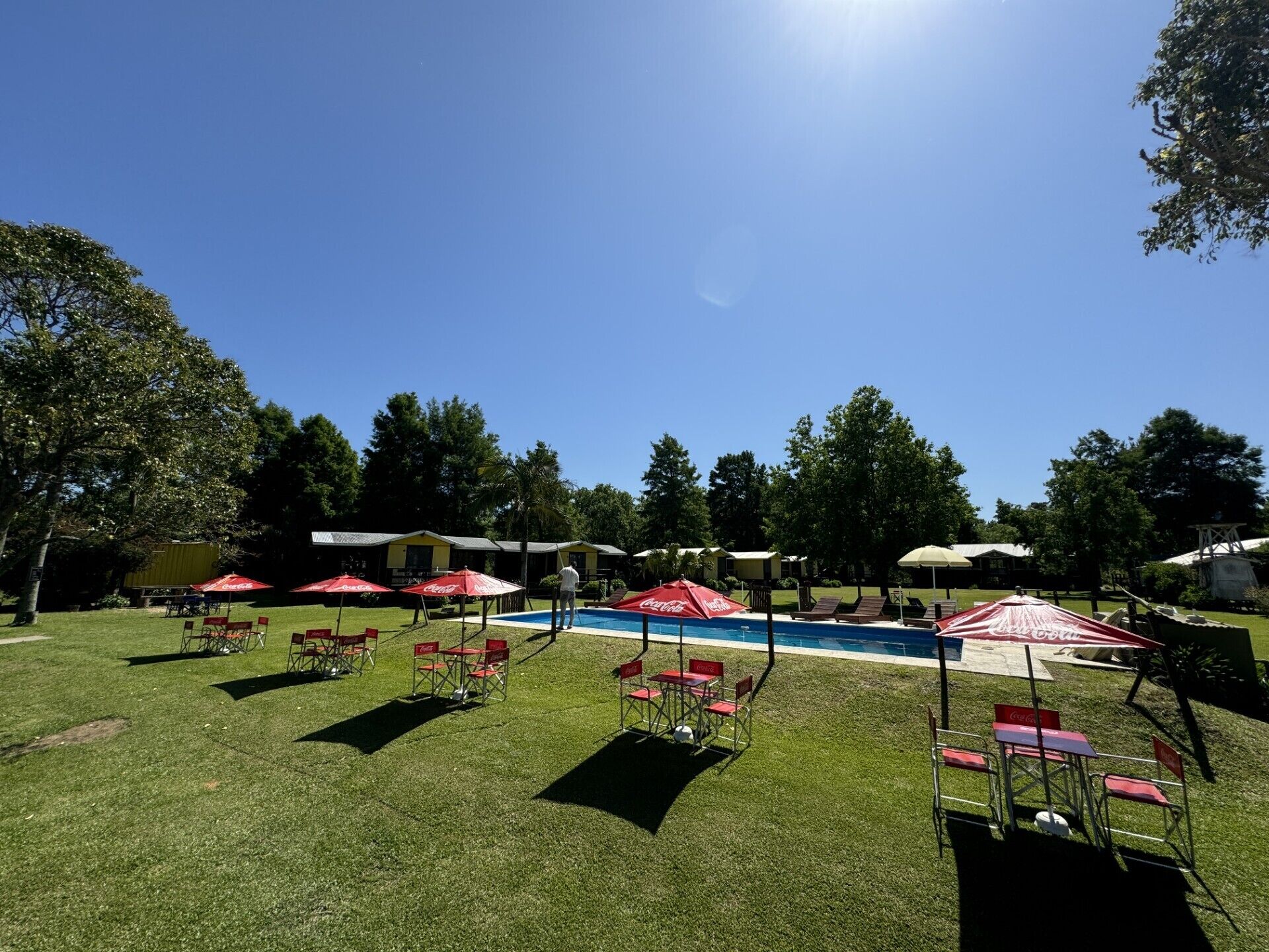 Un espace piscine agrémenté de chaises longues et de parasols, entouré de pelouse et d’arbres.