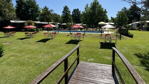 Zona de piscina con sillas de descanso y sombrillas, un muelle de madera al frente y un cielo azul despejado.